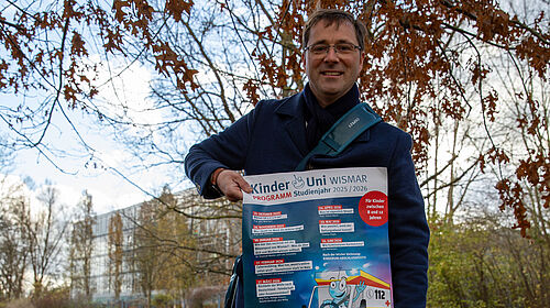KinderUni Hillmann Professor Hillmann steht auf einer Wiese, die mit bunten Blättern zugedeckt ist. In den Händern hält er ein Plakat. Im Hintergrund sind Gebäude der Hochschule zu sehen.