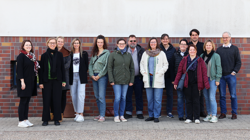 Gruppenfoto von 15 Personen, die in einer Reihe vor einer rot-braun geklinkerten Wand stehen. Alle blicken in die Kamera und lächeln. Die Kleidung ist lässig bis halbformell, die Stimmung freundlich und offen. Es handelt sich um Teilnehmende eines Zertifizierungsseminars.