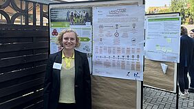 Eine blonde Frau mit Locken steht vor einem Poster. Sie trägt Businesskleidung. Auf dem Poster ist viel Schrift. Die Überschrift lautet LandStark.
