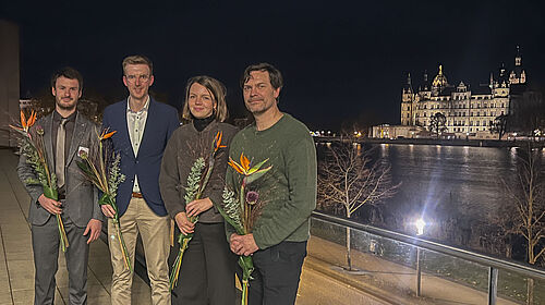 Die vier Personen stehen im Dunkeln auf einer Terasse. Im Hintergrund ist das angestrahlte Schweriner Schloss zu sehen.
