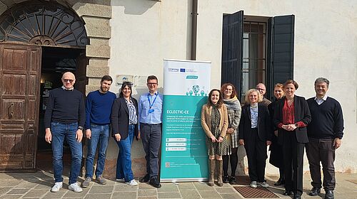 Gruppe von Projektpartnerinnen und -partnern steht vor einem historischen Gebäude neben einem Roll-up des Interreg-Projekts ECLECTIC-CE und blickt in die Kamera.