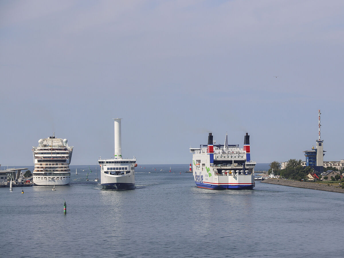 Three large ships are at the entrance to the port of Rostock-Warnemünde. A cruise ship is anchored on the left, a modern ferry with a striking white Flettner rotor enters the harbor in the middle, while another ferry leaves the harbor on the right.