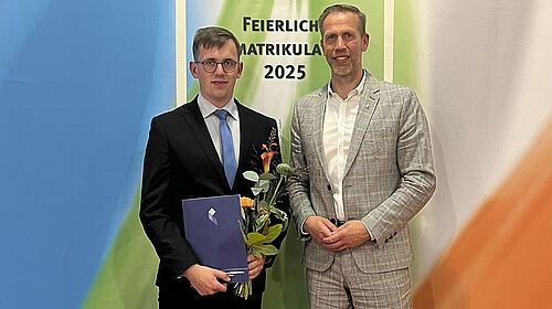 Die beiden Persoenn stehen vor farbigen Bannern, auf denen "Feierliche Immatrikulation 2025" steht. Der Ausgezeichnete hält Blumen und Urkunde in der Hand.
