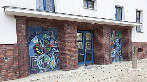 Ansichtsausschnitt der Fassade von Haus 18 mit Blick auf den Haupteingang. Das Eingangselement ist plastisch aus der weißen Putzfassade heraus gehoben. ine blaue Tür mit bunten Mosaiken links und rechts.