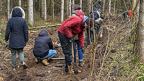 Es sind acht Personen im Wald zu sehen. Sie haben dicke Sachen an und graben mit Spaten Löcher für die einzusetzenden Pflanzen.