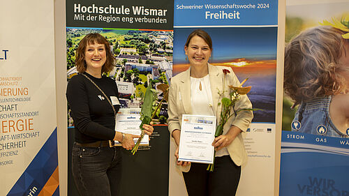 Die beiden jungen Frauen halten ihre Urunden sowie Blumensträuße in der Hand. Sie stehen vor Bannernder Schweriner Wissenschaftswoche und der Hochschule Wismar.