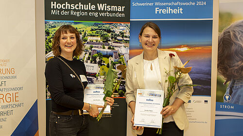 Die beiden jungen Frauen halten ihre Urunden sowie Blumensträuße in der Hand. Sie stehen vor Bannernder Schweriner Wissenschaftswoche und der Hochschule Wismar.