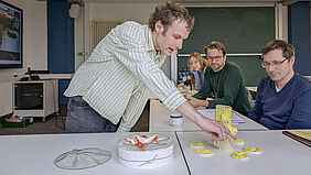 MMM-Präsentation Ein Student steht links von einem Tisch und zeigt auf eine gelbe Skulptur, die einer Wachskerze ähnelt. Rechts vom Tisch sitzen die beiden Professoren und schauen direkt auf die Skulptur.