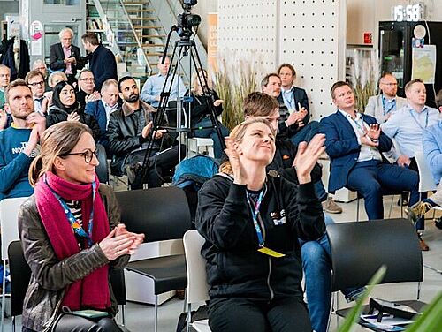 Ein vollbesetzter Veranstaltungsraum mit etwa 30-40 Personen, die aufmerksam einer Präsentation folgen. Die Teilnehmenden sitzen in mehreren Reihen auf schwarzen Stühlen. Im Raum sind professionelle Kameras auf Stativen aufgebaut. Die Atmosphäre ist konzentriert und professionell. Die Zuhörerschaft besteht aus Menschen verschiedenen Alters in Business-Casual-Kleidung. Die Beleuchtung ist hell und freundlich, und der moderne Veranstaltungsraum verfügt über technische Ausstattung für Präsentationen