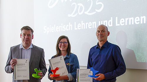 Lehrpreis2025 Die drei Personen sthen nebeneinander und halten ihre Urkunde sowie je einen Fischerpokal in Fakultätsfarbe in den Händen. Hinter Ihnen ist auf einem Bildschirm der Spruch "Spielerisch Lehren und Lernen" zu sehen.