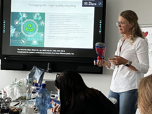 Eine Frau steht vor einem Bildschirm und hält eine Kunststoffverpackung in der Hand. Sie referiert zum Thema Recycling und Verpackungsgesetz. Auf dem Bildschirm ist eine Präsentationsfolie mit dem Titel „Packaging Act – high-quality recycling“ zu sehen. Auf dem Tisch vor ihr liegen verschiedene Verpackungen, Getränke und Notizen. Eine Teilnehmerin schreibt mit. Die Szene wirkt wie ein praxisnaher Workshop oder Vortrag zur Kreislaufwirtschaft.