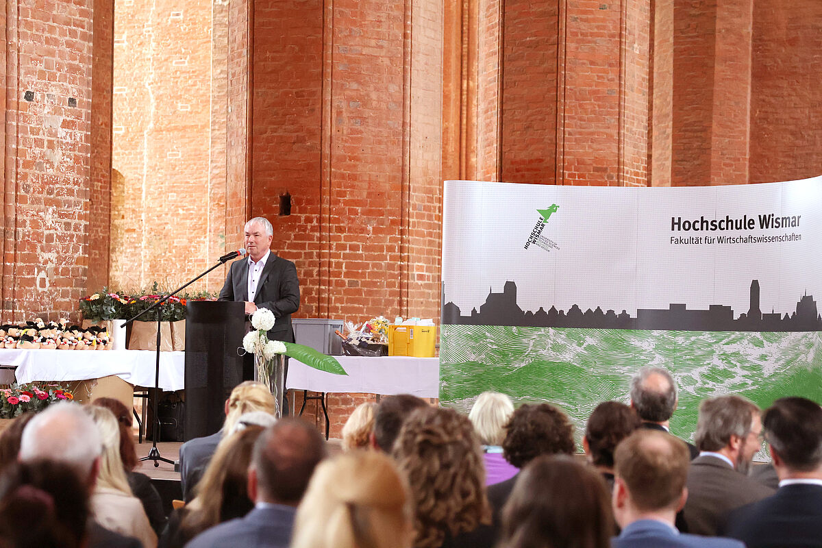 Der Bürgermeister der Hansestadt Wismar, Thomas Beyer auf der Rednerbühne vor dem Pult. Im Hintergrund der weiße Aufsteller der Fakultät Wirtschaftswissenschaften mit grünem Logo. Im Vordergrund ist erneut das sitzende Publikum zu erahnen.