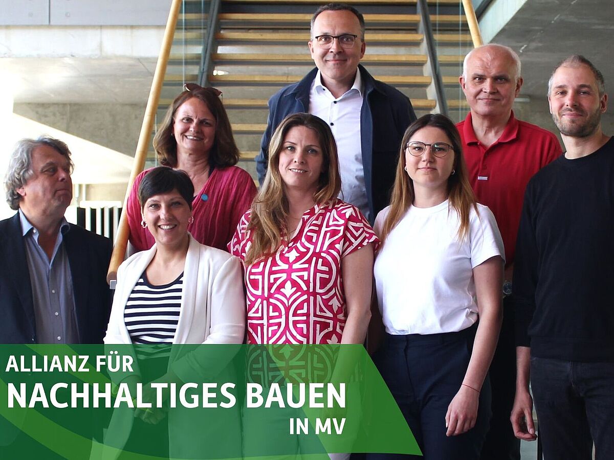 The picture shows a group of eight people posing in front of a staircase. In the background you can see a modern interior design with concrete walls and wooden railings.