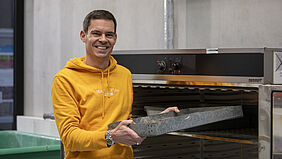 Prof. Hümme Der Professor steht vor einem Schrank, der wie ein großer Ofen aussieht. Er hält ein Blech in der Hand, das einem Backblech ähnlich sieht, und schiebt es in den Schrank.