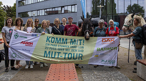 MUT-Tour2025 Zwei Pferde und mehrere Menschen stehen nebeneinander. Vor sich haben sie ein Banner ausgebreitet, auf dem die Aufforderung zur Teilnahme an der Mut-Tour steht: "Komm mit!" Außerdem sind die Logos der Unterstützereinrichtungen zu sehen.