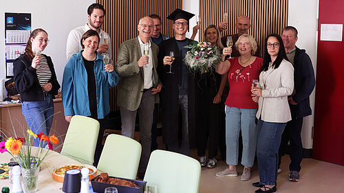 Celebration of Dominic Strube’s successful PhD. Dominic Strube stands amongst his colleagues following the successful completion of his PhD. The group is holding champagne glasses and celebrating the completion of his thesis. Dominic Strube is wearing a doctoral cap and holding a bouquet of flowers.
