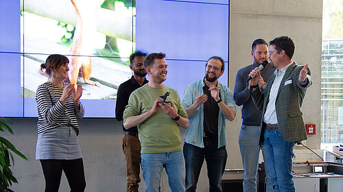 Science Slam 2025 Rechts unten am Bildrand sieht man die klatschenden Hände des Publikums. Auch die sechs Personen auf der Bühne klatschen, während der Sieger einen Fischer-Pokal in den Händen hält.