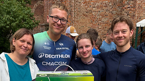 Eine Gruppe von vier Personen steht vor einer Backsteinmauer. Die zwei Frauen und zwei Männer lächelnd in die Kamera. Einer trägt erkennbar ein Fahrradtrikot