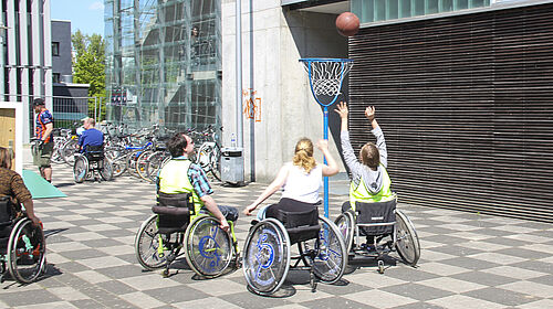 Drei Rollstuhlfahrende befinden sich vor einem niedrigem Basketballkorb. Eine der Personen wirft mit ausgestreckten Armen den Ball in Richtung Netz. Im Hintergrund sind Fahrräder vor dem Gebäude der Fakultät Gestaltung zu sehen.