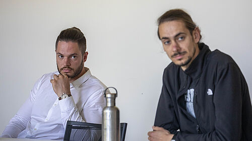 Das Bild zeigt zwei Männer an einem Tisch. Der linke trägt ein weißes Hemd, stützt seinen Kopf auf die Hand und blickt nachdenklich zur Kamera. Der rechte trägt eine schwarze Jacke, hat die Arme verschränkt und schaut leicht zur Seite.