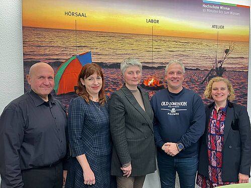 Fünf Personen stehen nebeneinander und lächeln in die Kamera. Im Hintergrund ist ein großformatiges Strandfoto bei Sonnenuntergang zu sehen, mit den Beschriftungen „Hörsaal“, „Labor“ und „Atelier“ sowie dem Hinweis „Hochschule Wismar – 10 Minuten bis zum Wasser“. Die Personen tragen eine Mischung aus formeller und legerer Kleidung.