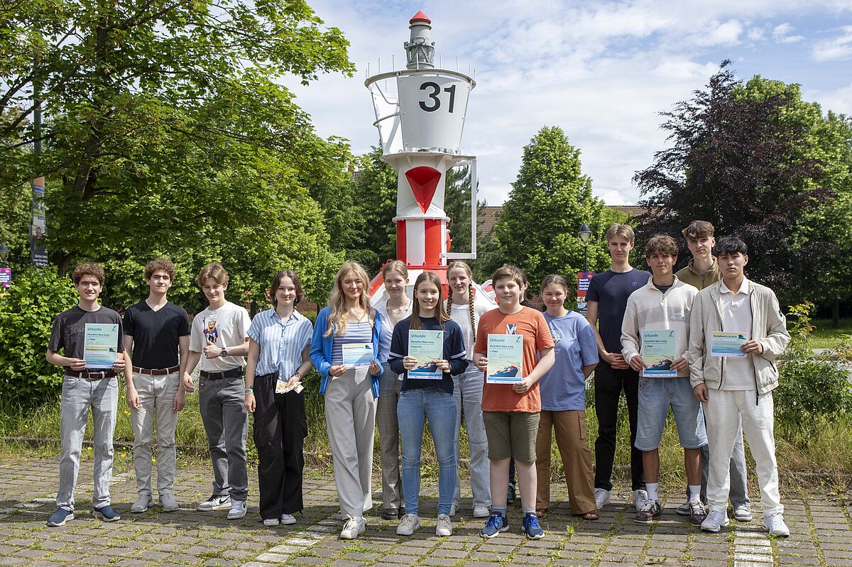 Die Gruppe steht draußen vor einem Seezeichen, einer rot-weiß-gestreiften Tonne, und hält Urkunden sowie Gutscheine in den Händen. Tonne