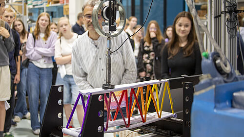 PBW25 Man sieht eine bunte Brücke aus Papier, die in eine MEtallvorrichtung eingespannt ist. Dahinter sind unscharf Schülerinnen zu erkennen, die zuschauen.
