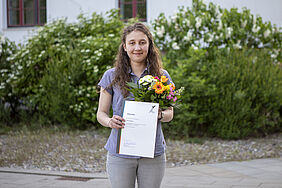 Die Studentin steh mit der Urkunde und einem Blumenstrauß in der Hand auf dem Campus.