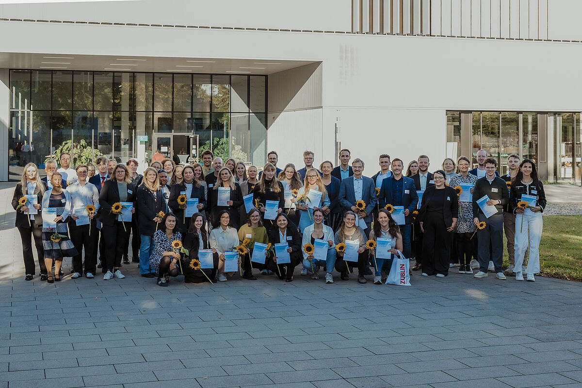StipendiatInnen und Förderer 2025/2026 50 Personen stehen neben- und hintereinander. Einige halten Blumen und Urkunden in den Händen. Daran erkennt man die Studierenden. Im Hintergrund ist Haus 6 zu erkennen.