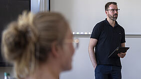 A man in a black polo shirt stands in front of a projection screen in a seminar room and holds a tablet in his hand. He is looking attentively in the direction of the audience. The blurred outline of a seated woman with blonde hair and glasses can be seen in the foreground.