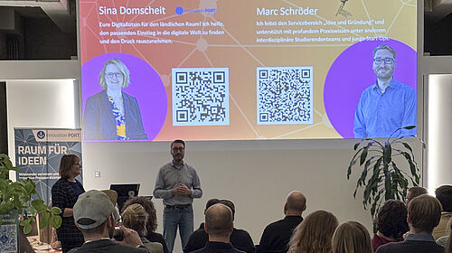 StartUpVoyage 2024 Man schaut über die Köpfe von 13 PErsonen hinweg auf eine Präsentation. Davor stehen Sina Domscheit und Marc Schröder.