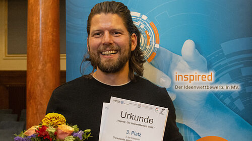 Ein Mann mit Bart und langem Haar hält lächelnd eine Urkunde für den 3. Platz bei einem Ideenwettbewerb in der Hand. Hinter ihm ist ein blaues Banner mit dem Schriftzug "inspired Der Ideenwettbewerb. In MV" zu sehen.