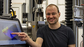 Mathias steht für einer Prüfmaschine, die viel höher als er ist und deshalb nicht auf´s Bild passt. Er zeigt auf den Bildschirm eines Laptops. Darauf ist ein farbiges 3D-Diagramm zu sehen.