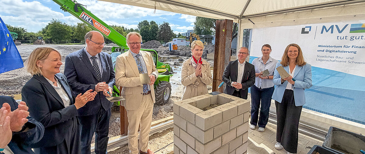 Several official representatives at the laying of the foundation stone for a new building, holding trowels, with MV and EU banners in the background