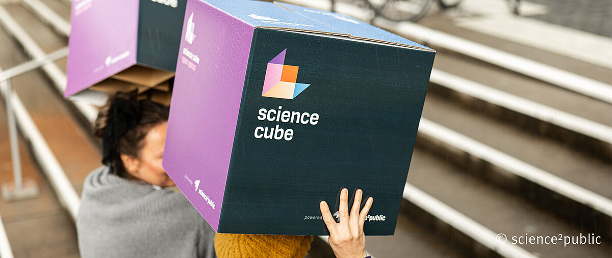 Zwei Personen tragen Papphocker mit der Aufschrift "Science Cube" eine Treppe hoch
