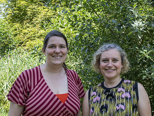 Zwei Frauen in Kleidern stehen lächelnd dem Betrachter frontal zugewandt. Im Hintergrund ist Wald zu sehen.