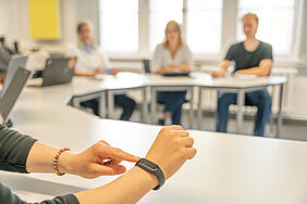 Im Vordergrund wird eine Smartwatch an einem Handgelenk bedient, während im Hintergrund unscharf vier Personen an Tischen in einem hellen Raum sitzen.