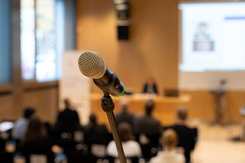 Ein Mikrofon im Vordergrund, scharfgestellt, mit einem unscharfen Konferenzraum im Hintergrund, in dem Personen einer Präsentation folgen.